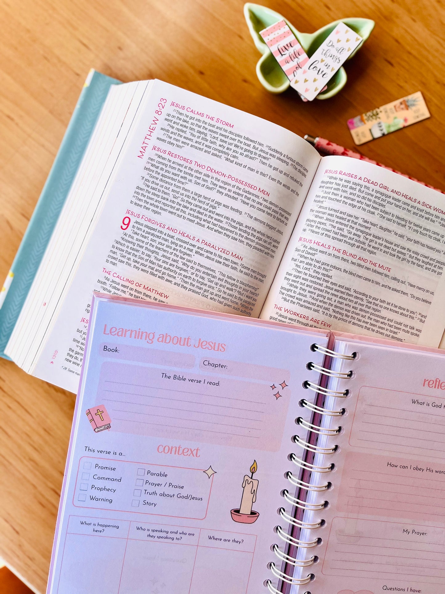 Open Bible and journal on a wooden surface with a small decorative item and bookmarks.