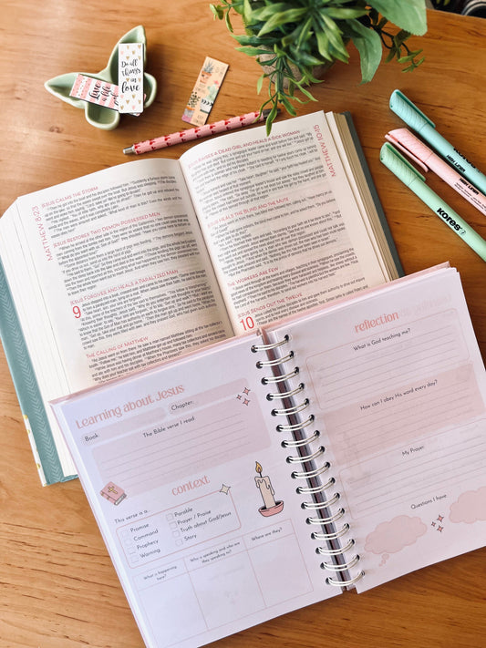 An open Bible with an open Teen Guided Prayer Journal on a wooden desk with a plant and stationery items. 