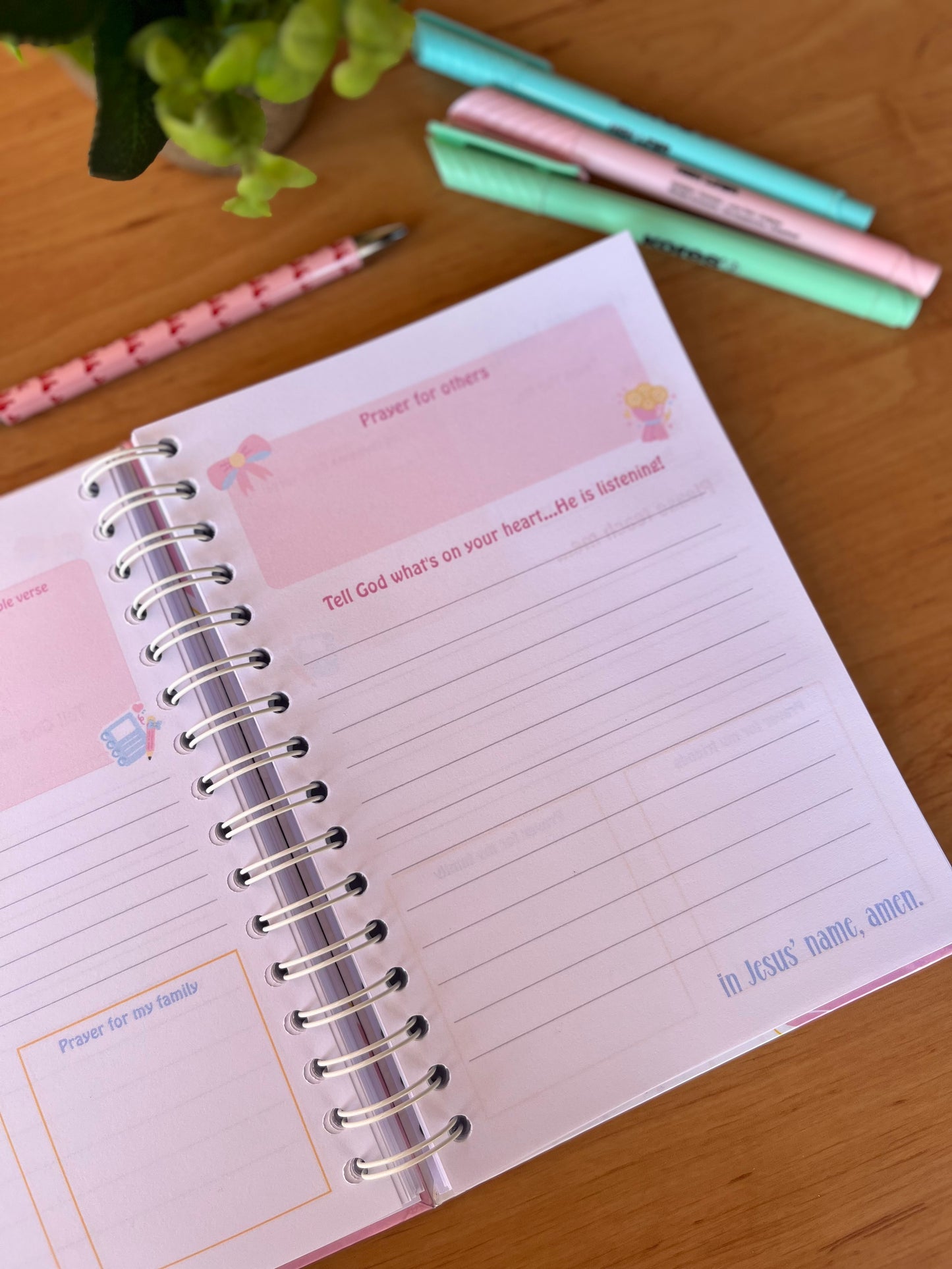 Open prayer journal pages on a wooden surface with pens and a plant.