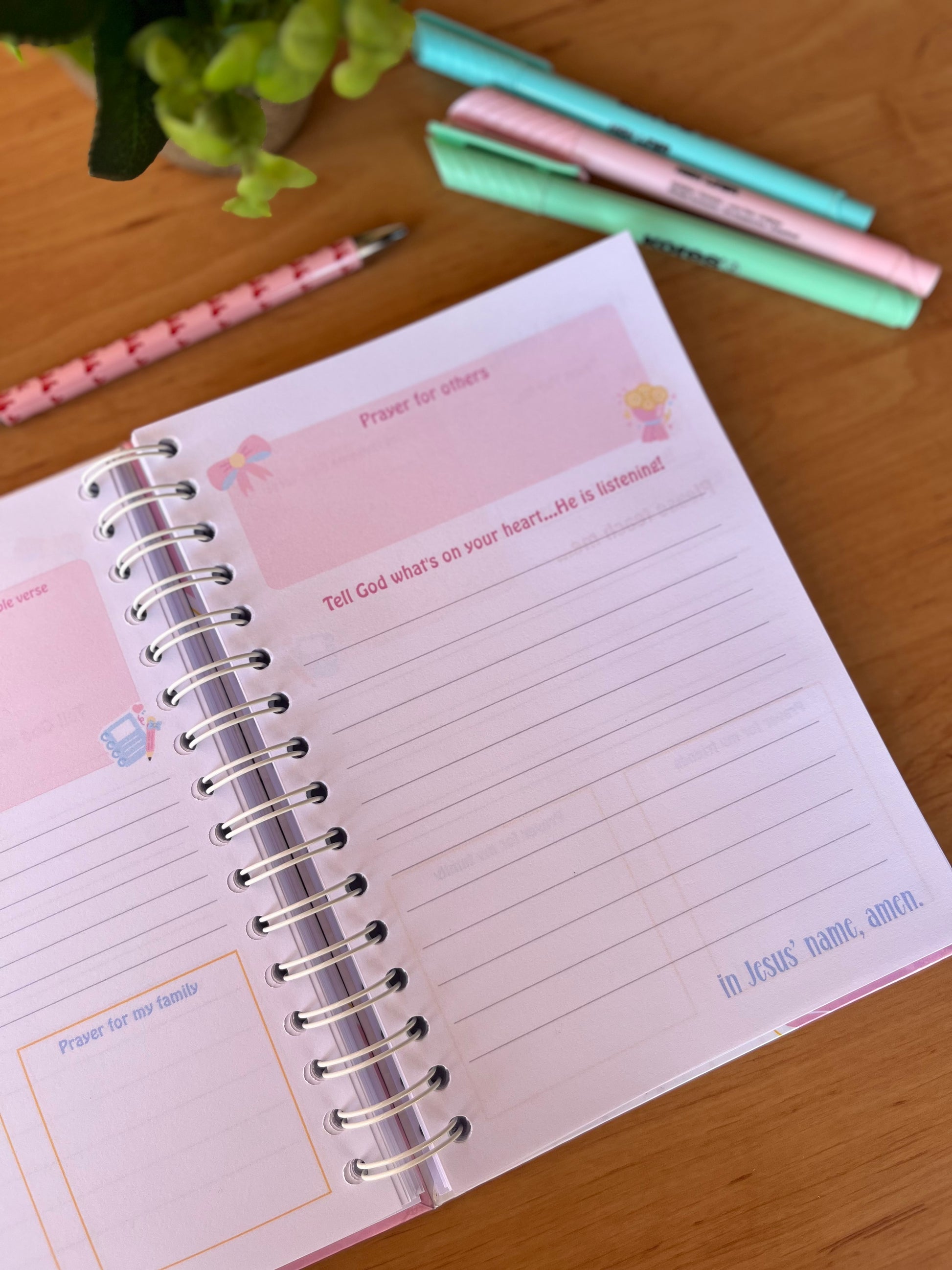 Open prayer journal pages on a wooden surface with pens and a plant.