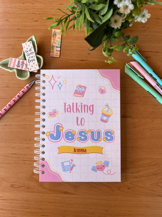 Notebook titled 'Talking to Jesus' on a wooden surface with stationery items and flowers.