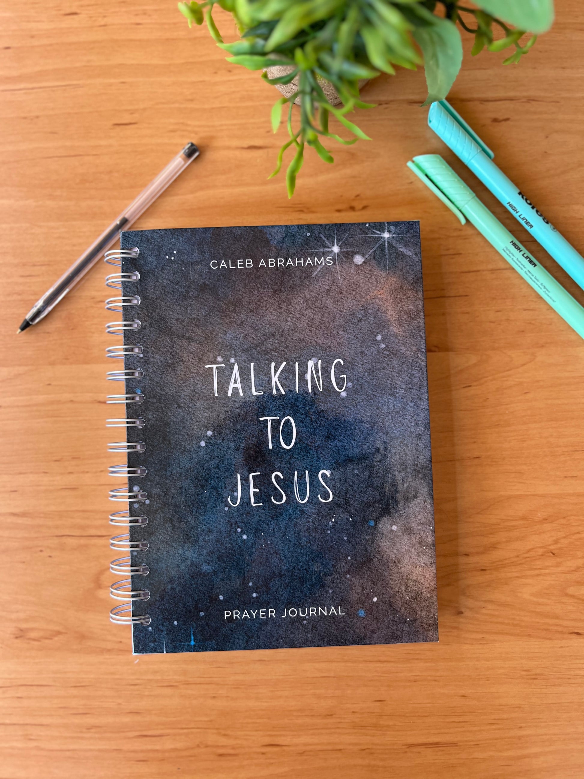 Prayer journal titled 'Talking to Jesus'  on a wooden surface with pens and a plant.