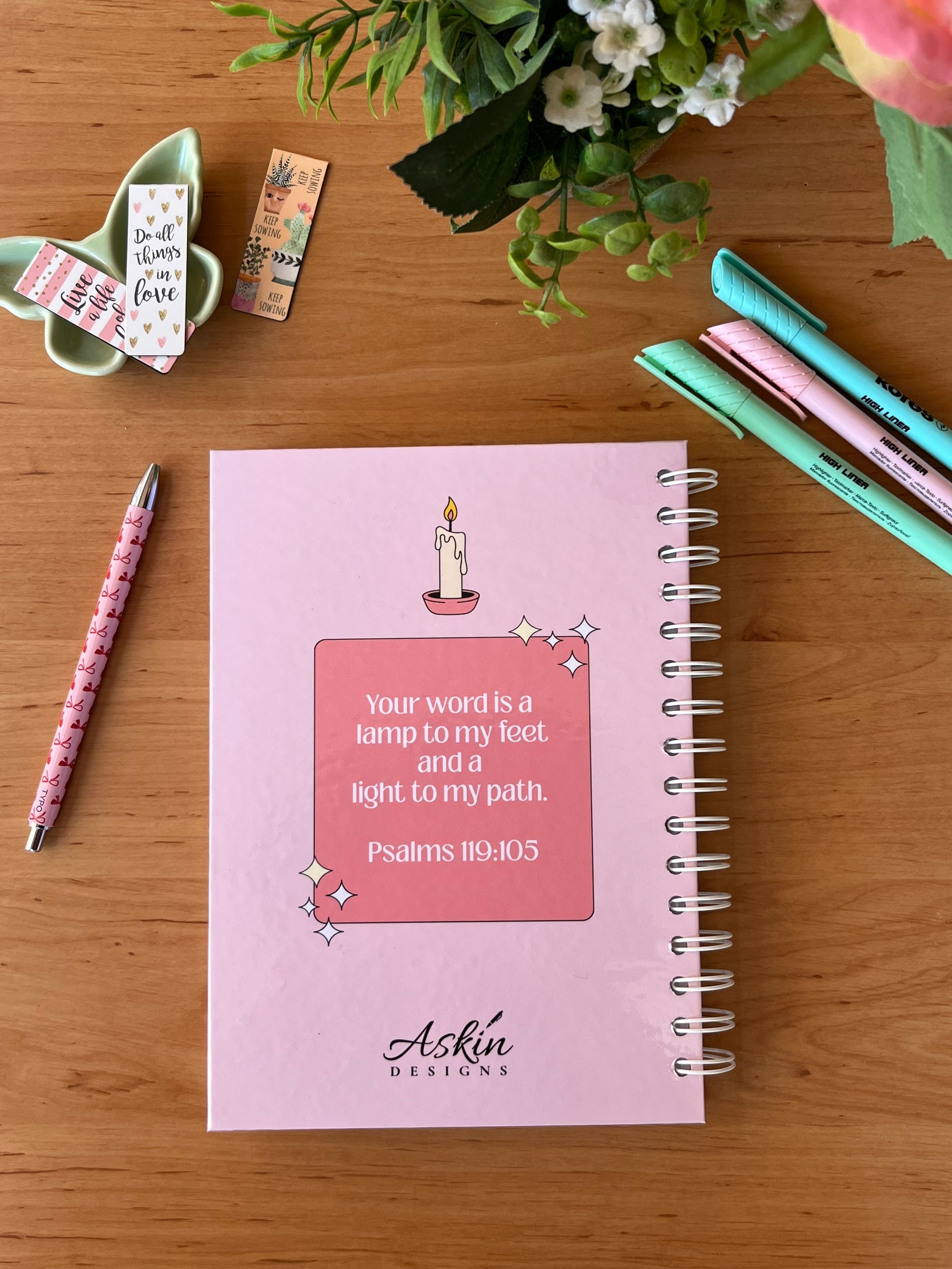 Pink notebook with Bible verse on a wooden surface with stationery items.