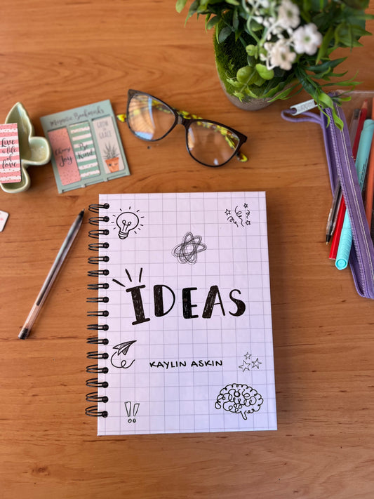 Notebook titled 'IDEAS' by Kaylin Askin on a wooden surface with stationery items.