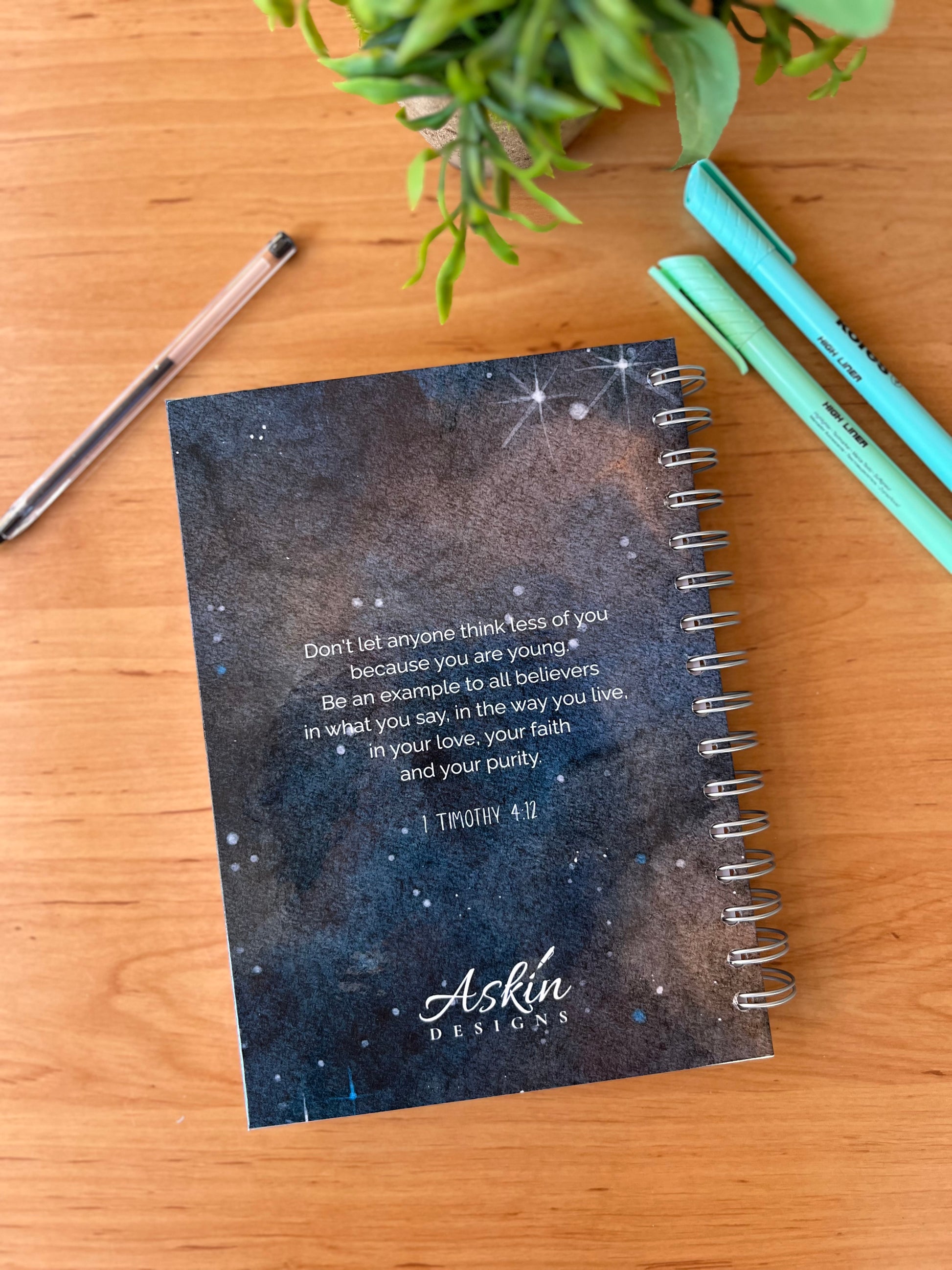 Notebook with Bible verse on a wooden surface with pens and a plant.